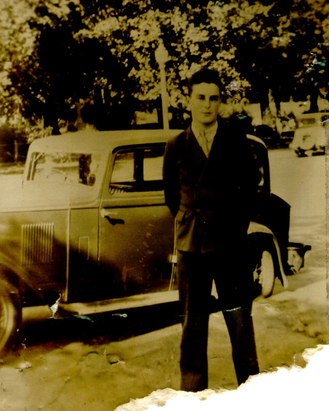 Damaged sepia photograph of a young man standing by a 1930s car, with creases, tears, and stains